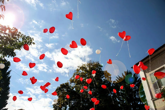 Many red heart-shaped balloons flying in a bright blue sky with white clouds and warm sunlight.