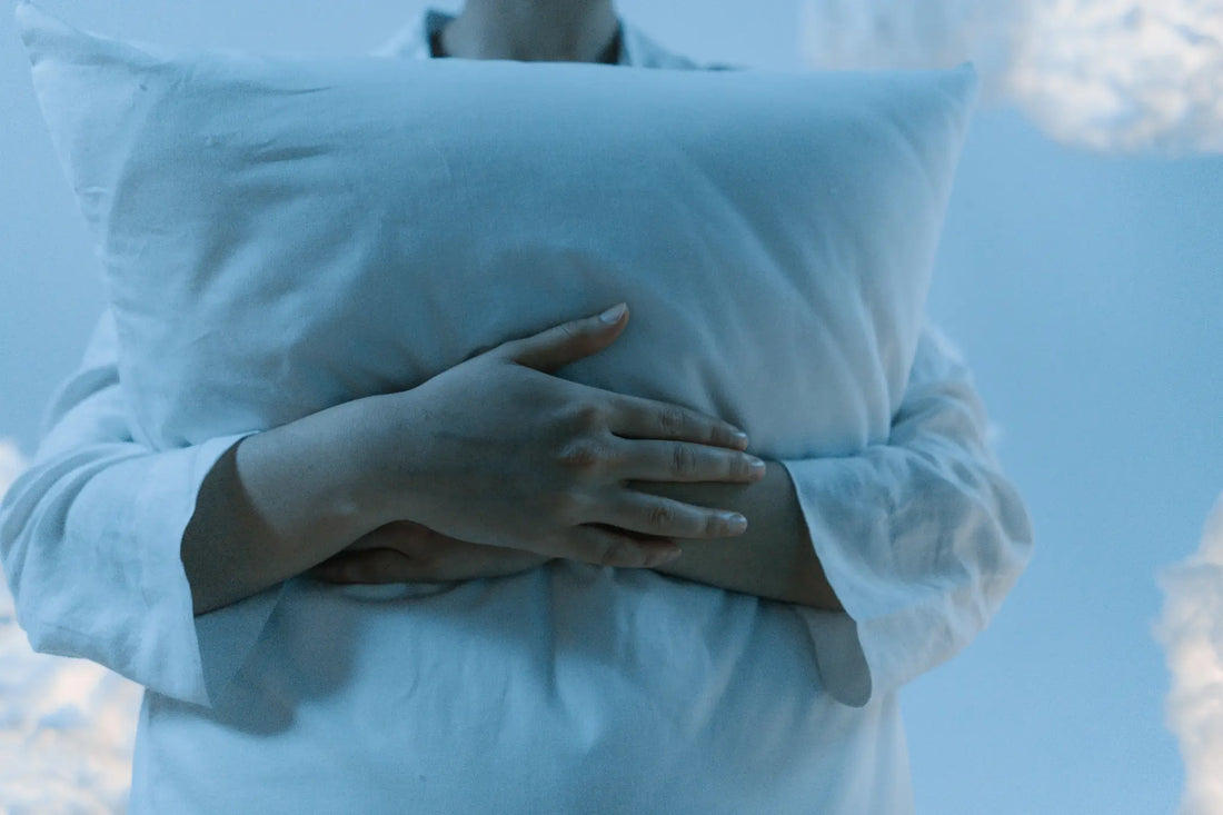 Person in soft pajamas hugging a light blue pillow against dreamy sky background
