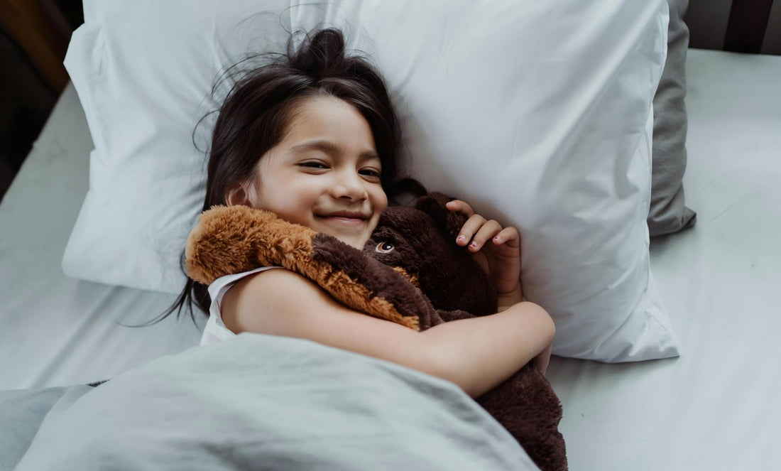Smiling little girl hugging fluffy brown bear plush toy on crisp white bed linens, soft calming companion for kids quiet rest sleep aid.