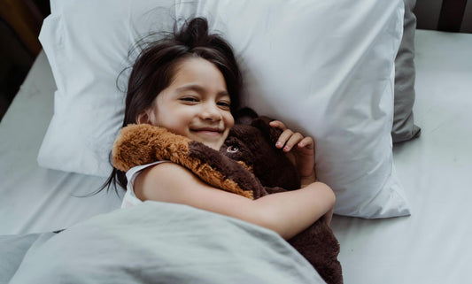 Smiling little girl hugging fluffy brown bear plush toy on crisp white bed linens, soft calming companion for kids quiet rest sleep aid.