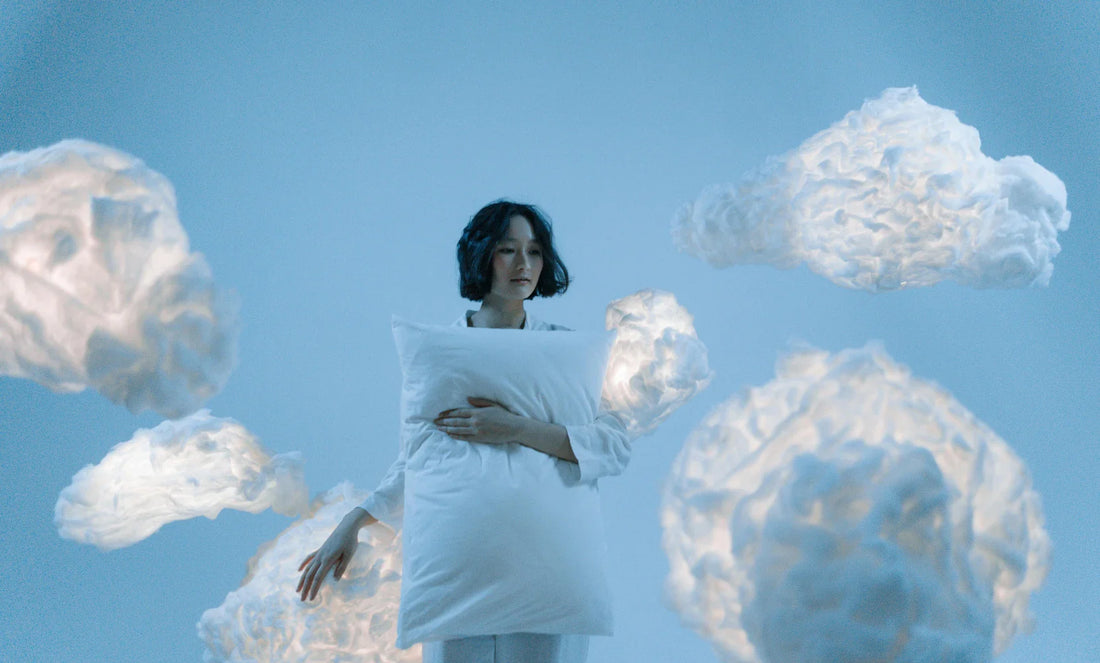 Woman in loungewear holding white pillow amid handmade cotton clouds on soft blue backdrop.