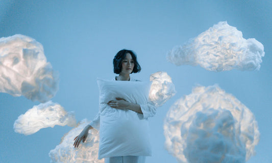Woman in loungewear holding white pillow amid handmade cotton clouds on soft blue backdrop.