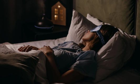 Woman sleeping with blue silk eye mask beside aroma humidifier on nightstand.